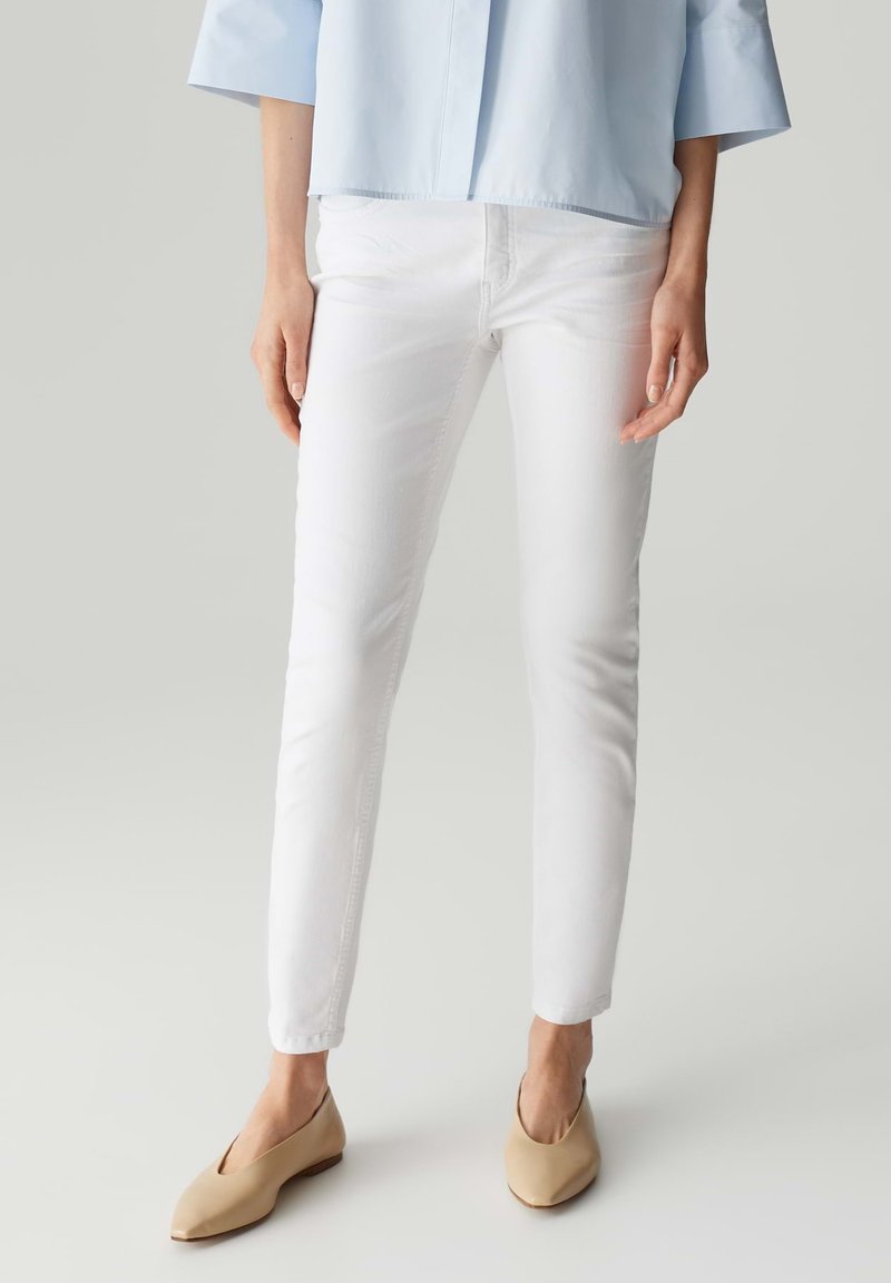 Jeans bianchi slim-fit con una texture liscia, abbinati a una camicia azzurro chiaro e ballerine beige con punta a forma appuntita.