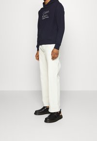 Felpa con cappuccio blu marina con un logo sul frontale, abbinata a pantaloni bianchi e scarpe slip-on nere con finitura testurizzata e suole a contrasto.