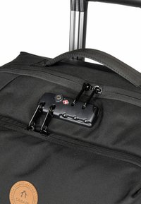 Sac à dos en tissu noir avec une poche zippée, des accessoires noirs et un patch rond beige. Équipé d'un mécanisme de verrouillage pour une fermeture sécurisée.