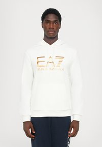 Moletom branco com bolso canguru, apresentando um logo EA7 em metalizado dourado ao longo do peito. Tecido suave e texturizado, corte descontraído.