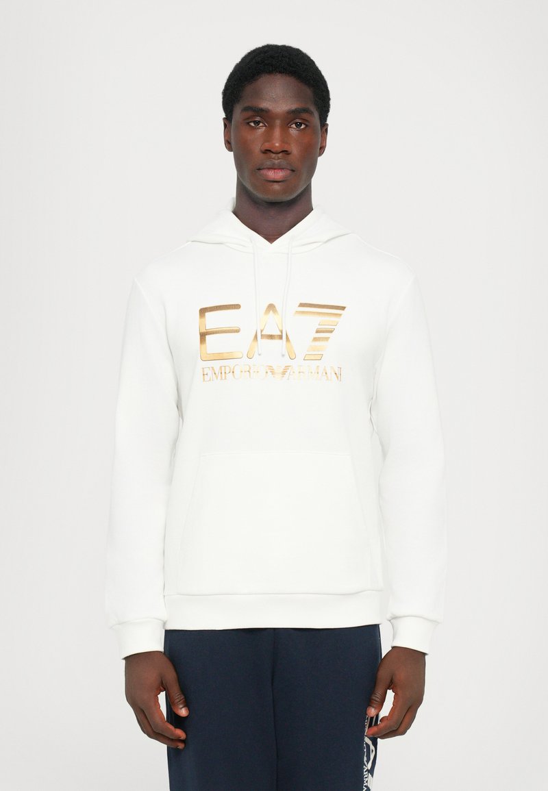 Moletom branco com bolso canguru, apresentando um logo EA7 em metalizado dourado ao longo do peito. Tecido suave e texturizado, corte descontraído.