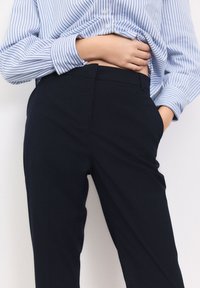 Pantalon bleu marine sur mesure avec un design élégant, comportant une ceinture plate et des poches, associé à une chemise boutonnée rayée bleu clair.
