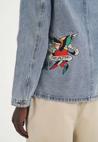 Vaalean sininen farkkutakki, jossa on brodeerattu sydän, kotka ja tekstisuunnittelu takana, hopeiset nappikoristeet ja erottuva ompelu.