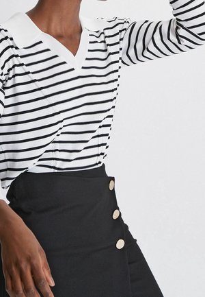 Personne portant une chemise blanche à manches longues avec des rayures noires et un col, associée à une jupe noire taille haute ornée de trois boutons dorés.