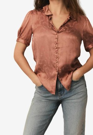 Femme portant un chemisier boutonné à manches courtes, de couleur rouille brillante avec un motif floral subtil, et un jean taille haute bleu clair, les mains dans les poches.