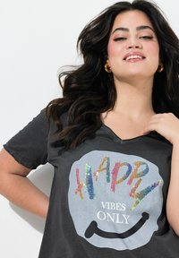 Donkergrijze T-shirt met een V-hals, voorzien van een kleurrijke, sequined "HAPPY"-graphic en een smiley-gezicht ontwerp. Zachte, katoenachtige structuur.