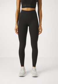 Høyt liv svarte leggings laget av elastisk materiale, med en jevn tekstur og en figurnær design, sammen med grå og rosa joggesko.