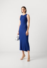 Robe bleue à côtes avec design sans manches et accents de boutons en argent. Le mannequin tient un petit sac argenté, portant des talons beiges à lanières.