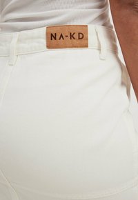Jean en denim blanc avec une finition texturée, arborant une étiquette en cuir marron embossée avec "NA-KD" sur la taille.