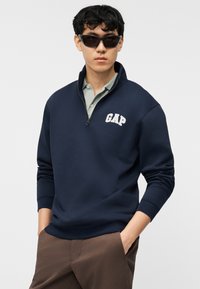 Νεαρός άνδρας με σκούρα γυαλιά ηλίου, μπλε ναυτικό φούτερ GAP με φερμουάρ έως τη μέση πάνω από ανοιχτό γκρι πόλο και καφέ παντελόνια, έχοντας τα χέρια στις τσέπες.