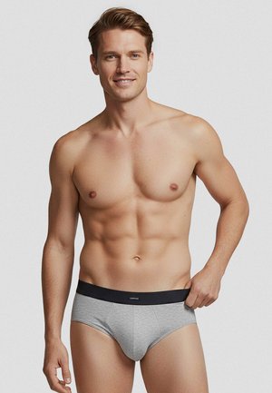 Smilende mand uden skjorte iført grå underbukser med sort taljebånd, stående mod en ensfarvet lys baggrund, hånd trækker let i taljebåndet.