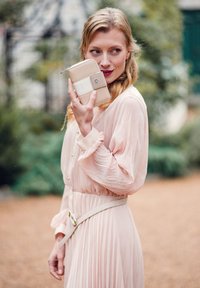 Beige peněženka s hnědým popruhem, zipovým uzávěrem a kovovým logem. Nosená k jemně růžovým skládacím šatům se staženým pasem a detaily na knoflíky.