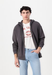 Mørkegrå zip-up hettegenser med hette, brukt over en hvit grafisk T-skjorte og lyseblå jeans. Glatt tekstur og avslappet passform.
