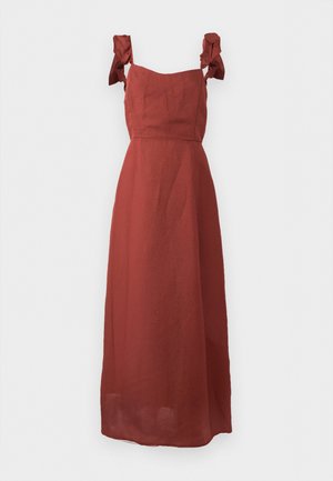 Μάξι φόρεμα χωρίς μανίκια σε χρώμα σκουριάς με τετράγωνο ντεκολτέ και βολάν στα τιραντάκια σε μονόχρωμο φόντο.