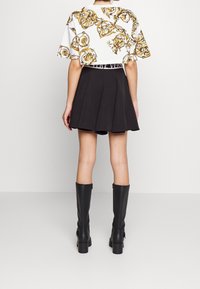 Bílý crop top se zlatým barokním vzorem, spárovaný s černou rozšířenou mini sukni. Modelka má na sobě černé kozačky na vysokém podpatku až ke kolenům.