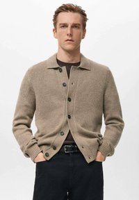 Beige gebreid vest met een wijde kraag, knoopsluiting aan de voorkant en geribbelde manchetten. Draagt over een donker T-shirt, gecombineerd met een donkere broek.