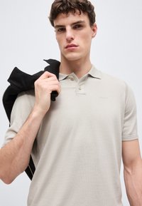 Jeune homme aux cheveux bruns courts portant un polo beige clair, tenant une veste noire sur son épaule sur un fond uni.