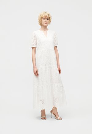 Γυναίκα με κοντά σγουρά ξανθά μαλλιά φοράει ένα μακρύ λευκό φόρεμα με κεντήματα και μανίκια φουσκωτά, καθώς και μπεζ ψηλοτάκουνα σανδάλια, στέκεται μπροστά σε λευκό φόντο.