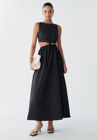 Model poseert in een zwart uitgesneden jurk met een strik taille, terwijl ze een crèmekleurige clutch vasthoudt. Ze draagt zwarte sandalen en heeft elegante oorbellen.