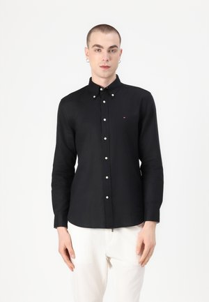 Camicia nera a maniche lunghe con bottoni, colletto, bottoni bianchi e un logo sottile sul petto, abbinata a pantaloni chiari.
