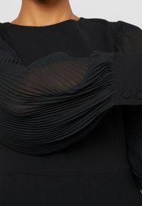 Schwarzes Kleid mit strukturierten, gefältelten, durchsichtigen langen Ärmeln und geknöpften Manschetten, das einen glatten, figurbetonten Oberkörper und einen runden Ausschnitt aufweist.