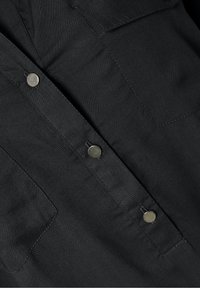 Tissu noir avec deux poches poitrine et boutons-pression métalliques au centre, montrant un gros plan des détails et coutures à l'avant de la chemise.