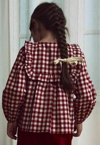 Blusa a cuadros rojos y blancos con cuello y mangas con volantes, presenta un lazo en la parte de atrás, fabricada con un tejido ligero.