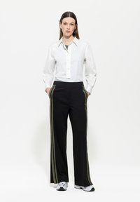 Weißes Hemd mit langen Ärmeln, kombiniert mit schwarzen Hosen, die grüne Streifen an den Seiten haben. Das Model trägt graue Sneakers.