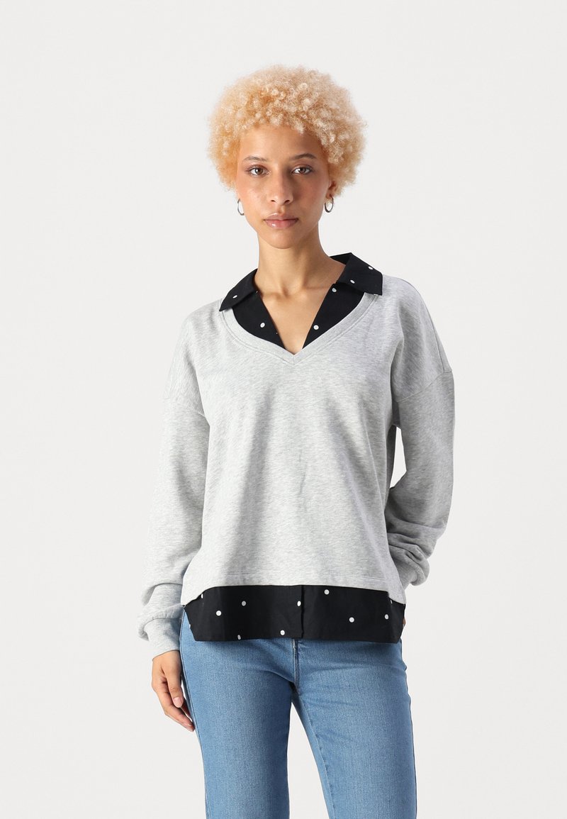 Grå hettegenser med V-hals, sort prikkete skjortebunn, lange ermer og løs passform. Blå jeans fullfører antrekket.
