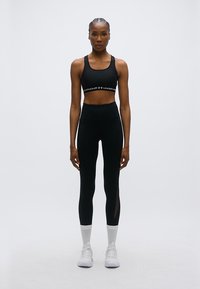 Μαύρο αθλητικό κοντό τοπ με λωρίδα λογότυπου και κολάν ψηλόμεσα με πλαϊνή πλέξη από δίχτυ, σε συνδυασμό με λευκά αθλητικά παπούτσια χαμηλού ύψους και κάλτσες.