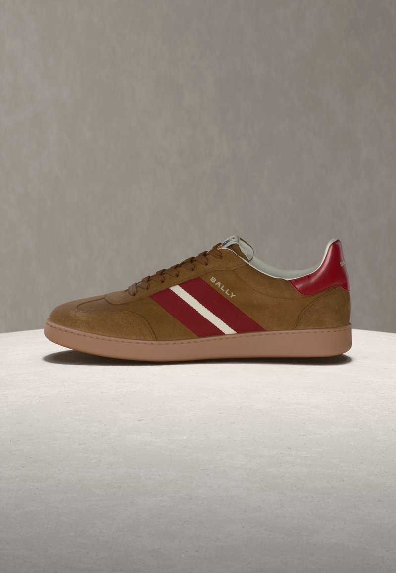 Καφέ σουέντ αθλητικό παπούτσι με κόκκινες και λευκές ρίγες, κόκκινη γλώσσα στη φτέρνα και επίπεδη σόλα από καουτσούκ, με την επωνυμία "Bally" στο πλάι.