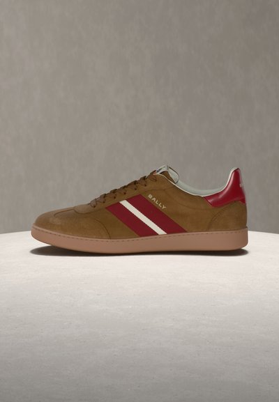 Barna velúr tornacipő piros és fehér csíkokkal, piros sarokfüllel és lapos gumitalppal, az oldalán "Bally" márkajelzéssel.