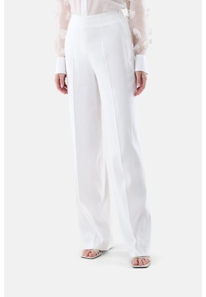 Frau trägt hoch taillierte weiße weit geschnittene Hose und eine transparente weiße Langarmbluse mit floralen Details, stehend auf weißem Hintergrund.