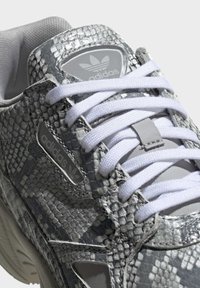 Grå Adidas-sneakers har ett texturerat ormskinnsmönster, vita snören och en plastlogotypdetalj på tungan. Tjock såldesign.