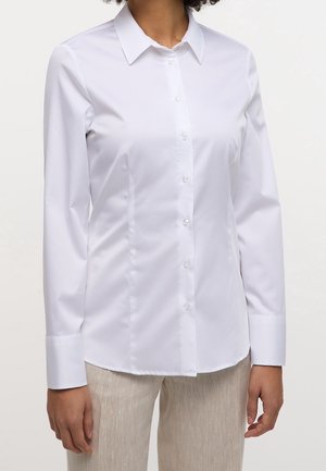 Personne portant une chemise blanche ajustée à manches longues boutonnée et un pantalon beige clair, montrée des épaules aux hanches sur un fond uni.