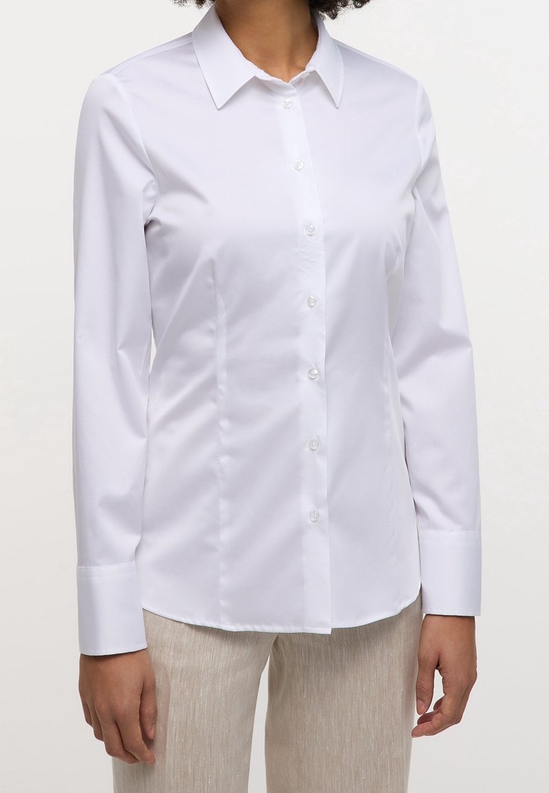 Personne portant une chemise blanche ajustée à manches longues boutonnée et un pantalon beige clair, montrée des épaules aux hanches sur un fond uni.