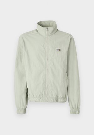 Lys grønn zip-up jakke med høy krage, samlede mansjetter, elastisk midje og et lite logo-patch på venstre bryst.