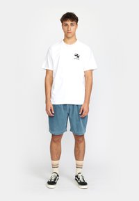 Witte katoenen t-shirt met een zwart graphic op de linkerkant van de borst, gecombineerd met blauwe corduroy shorts, gestreepte sokken en zwarte sneakers.