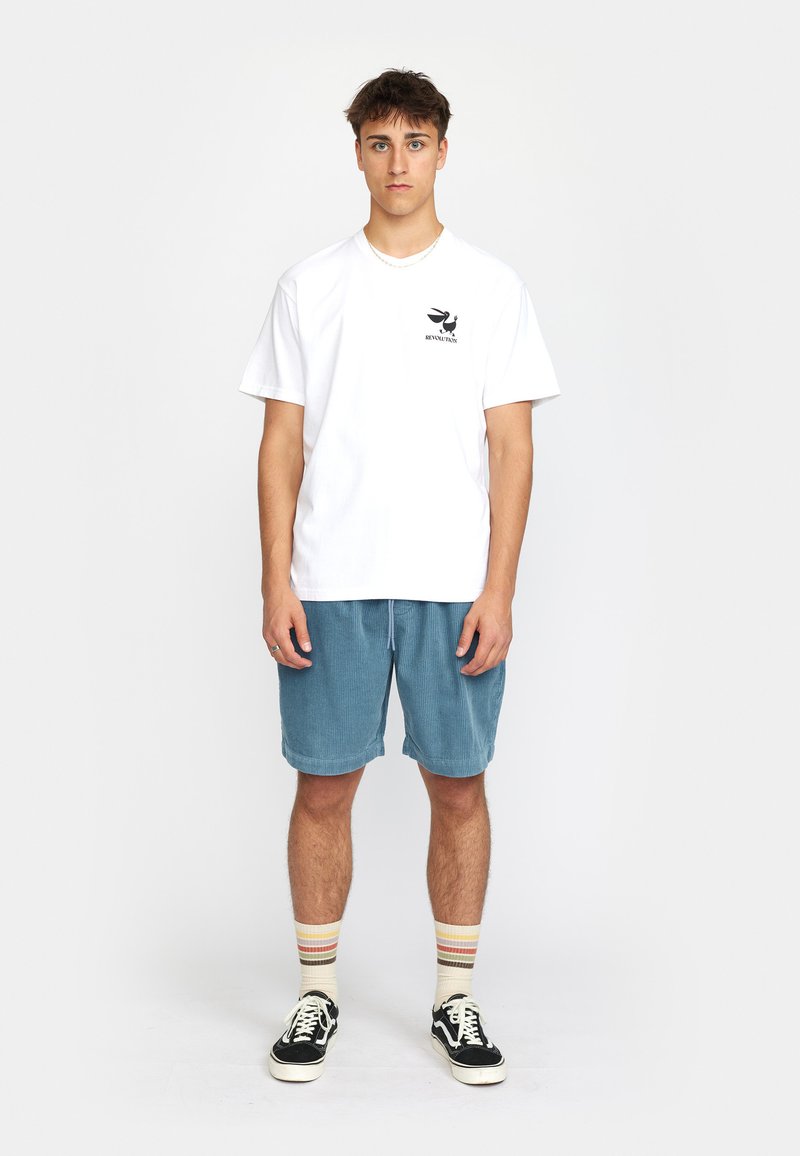 Witte katoenen t-shirt met een zwart graphic op de linkerkant van de borst, gecombineerd met blauwe corduroy shorts, gestreepte sokken en zwarte sneakers.