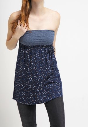 Vrouw met lang rood haar draagt een strapless donkerblauwe top met kleine witte en roze stippen, gecombineerd met een zwarte broek, tegen een witte achtergrond.