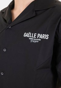 Camicia nera con colletto a punta, con testo bianco "GAËLLE PARIS HOMME COLLECTION" sul lato sinistro del petto. Tessuto liscio, apertura con bottoni.