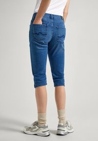 Džínové capri kalhoty ve středně modré barvě, se dvěma zadními kapsami s obloukovým prošíváním, ukončené pod kolenem, doplněné šedými teniskami a krémovými ponožkami.