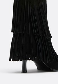 Stivaletto nero con tacco alto, decorato con frange, realizzato in materiale morbido, con una superficie liscia e un tacco sottile e sagomato.