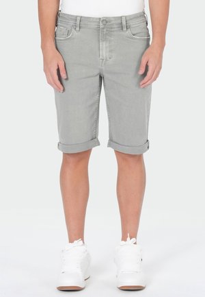 Grau aufgerollte Jeansshorts, getragen von einer Person in weißen Sneakers, von der Taille bis zu den Füßen vor neutralem Hintergrund gezeigt.