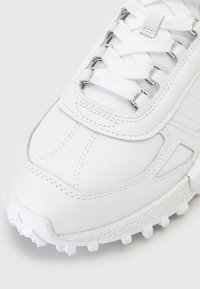 Gros plan sur l'avant d'une basket blanche avec une semelle texturée, une tige en cuir cousu et des lacets blancs passés dans des œillets en métal.