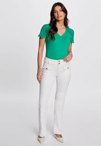 Top verde con escote en pico y detalle de encaje en las mangas, combinado con pantalones blancos de cintura alta con bolsillos frontales y acentos de botones, llevados con tacones beige.