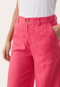 Pantaloni di lino rosa con vestibilità rilassata, dotati di tasche frontali e di un bottone in legno naturale in vita. Tessuto leggero e trama liscia.