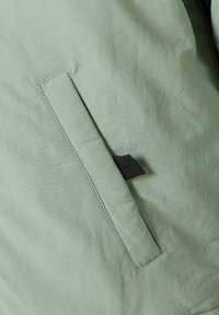 Veste en tissu vert clair avec une texture subtile, dotée d'une poche latérale et d'un onglet d'accent noir près de l'ouverture.