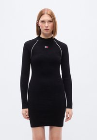Μαύρο ριμπ πλεκτό φόρεμα με μακριά μανίκια και ψηλό λαιμό. Διαθέτει λευκό διακοσμητικό ράψιμο και ένα μικρό λογότυπο στο στήθος.