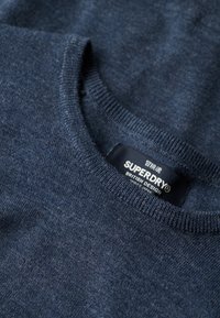 Marineblauwe crewneck trui van zachte, gestructureerde stof. Heeft een geribbelde kraag en een label met "SUPERDRY" branding.